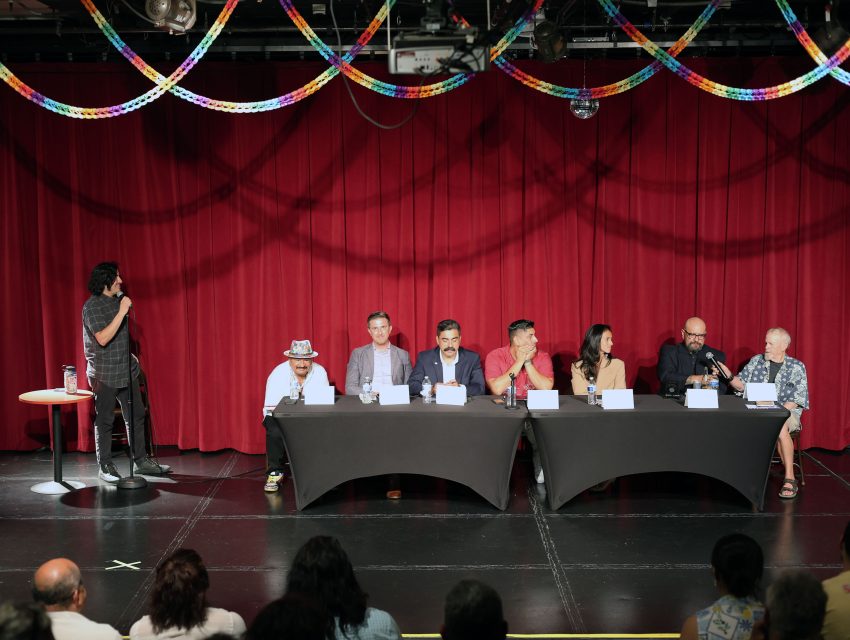 Seven people sit at a table on a stage with a red curtain backdrop. One person stands to the side with a microphone, addressing the panel. A decorated ceiling and audience are visible.