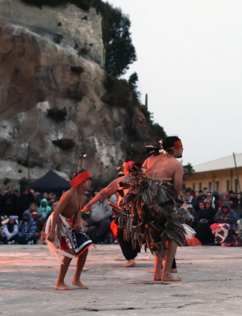People in traditional attire perform a cultural dance outdoors, with a rocky cliff in the background and a seated audience watching.