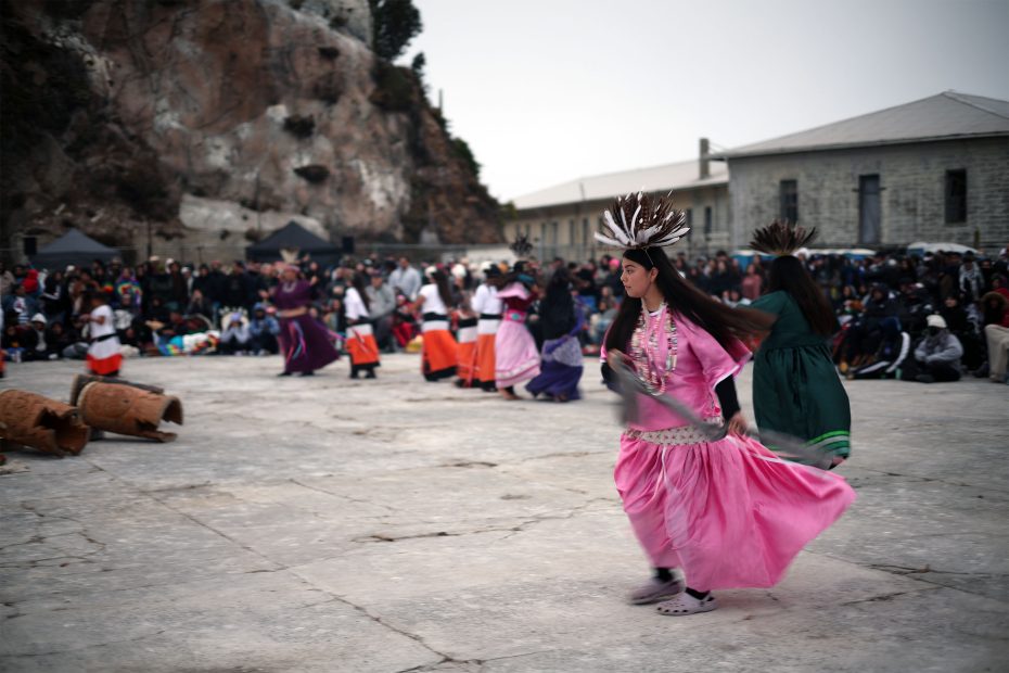 People in colorful traditional attire dance in a large outdoor area, with a crowd watching and rocky landscape in the background.