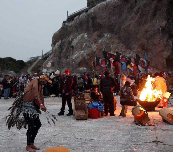 A group of people gather outdoors near a large fire, with some wearing traditional attire and others watching. Flags are displayed in the background.