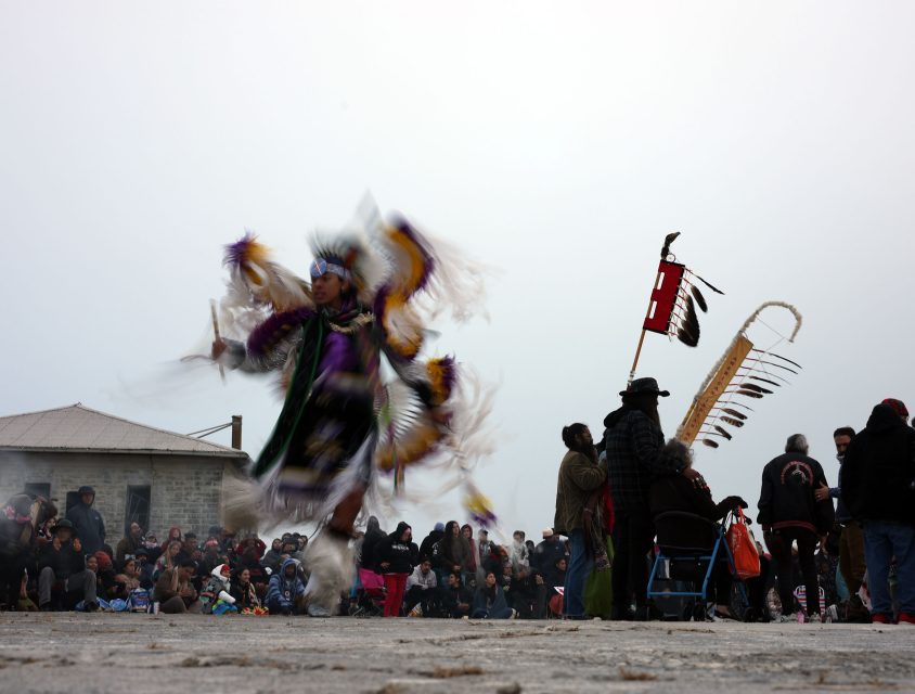 A dancer in traditional attire performs at an outdoor event, with a crowd watching in the background.