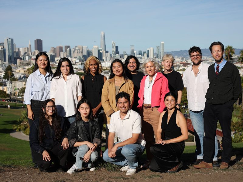 A group of people posing outdoors with a city skyline in the background on a sunny day.