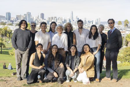 A group of people posing outdoors with a city skyline in the background on a grassy area. Some are seated and others standing. It's a sunny day.
