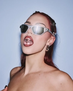 A person wearing futuristic sunglasses and red lipstick shows off metallic tooth jewelry against a light blue background.