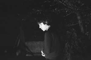 A person stands outdoors at night, partially illuminated, with trees and shadows in the background.