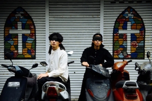 Two individuals sit on scooters in front of a wall with stained glass cross designs.