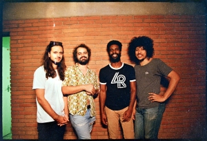 Four men standing together in front of a brick wall, wearing casual clothing.