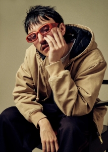 A person wearing red glasses and a tan hoodie sits with hand on face against a neutral background.