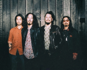 Four men stand against a dark wooden wall, each wearing casual clothing. One wears sunglasses. They have long hair and relaxed expressions.