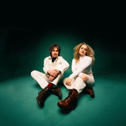 Two people sitting on a teal floor, both wearing white outfits and brown boots, against a dark background.