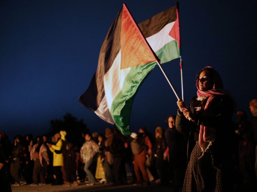 A person holds a Palestinian flag at a nighttime gathering with a group of people in the background.