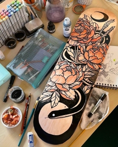 A partially painted skateboard deck featuring a floral design with peonies and a snake is surrounded by art supplies, including markers, paint bottles, brushes, and a sketchbook on a cluttered workspace.