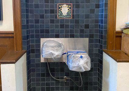 Drinking fountains covered with plastic bags and tape, set against a dark tiled wall with a decorative owl tile above.