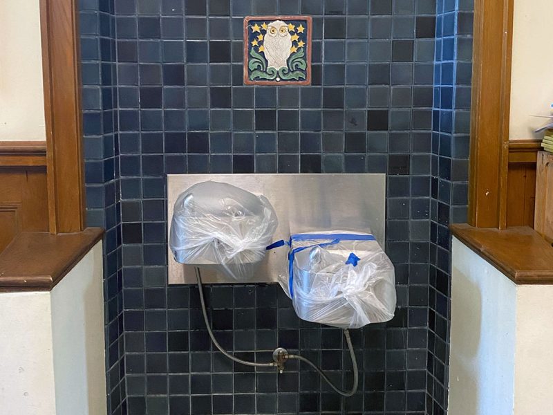 Drinking fountains covered with plastic bags and tape, set against a dark tiled wall with a decorative owl tile above.