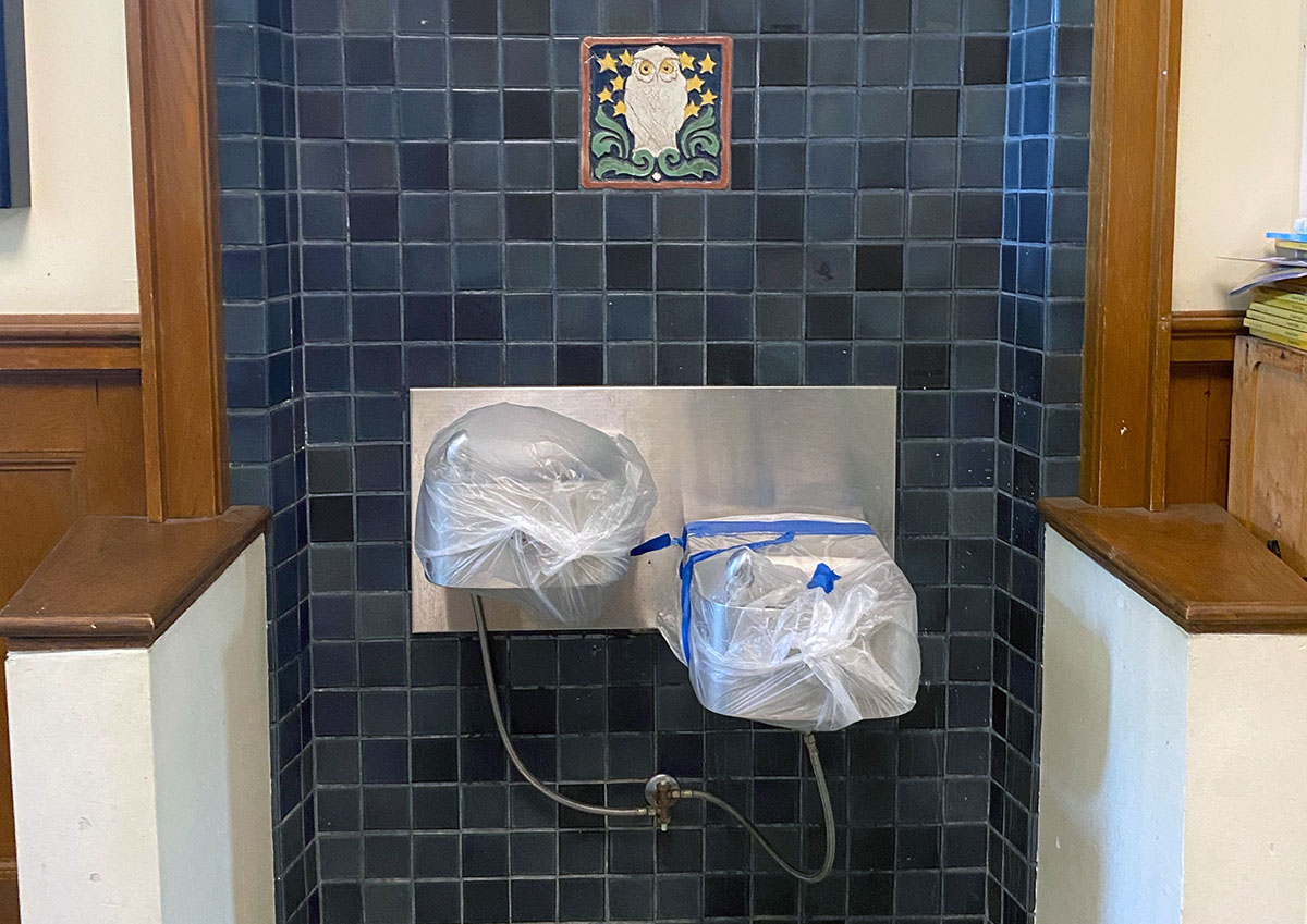 Drinking fountains covered with plastic bags and tape, set against a dark tiled wall with a decorative owl tile above.