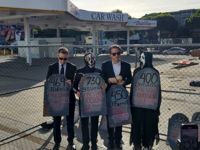 Four people in suits and masks stand holding signs resembling tombstones near a fenced construction site.