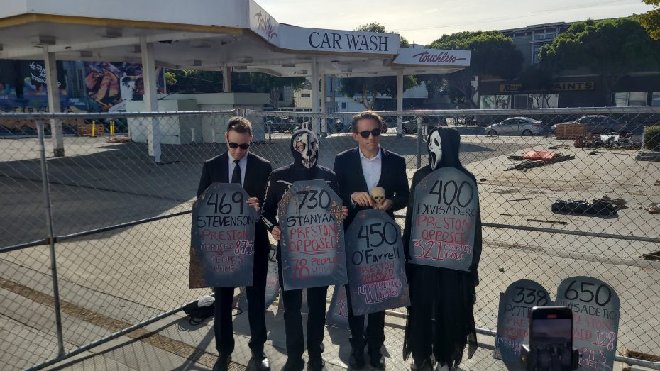 Four people dressed in black suits and masks holding signs in front of a fenced, closed gas station with a car wash sign in the background.