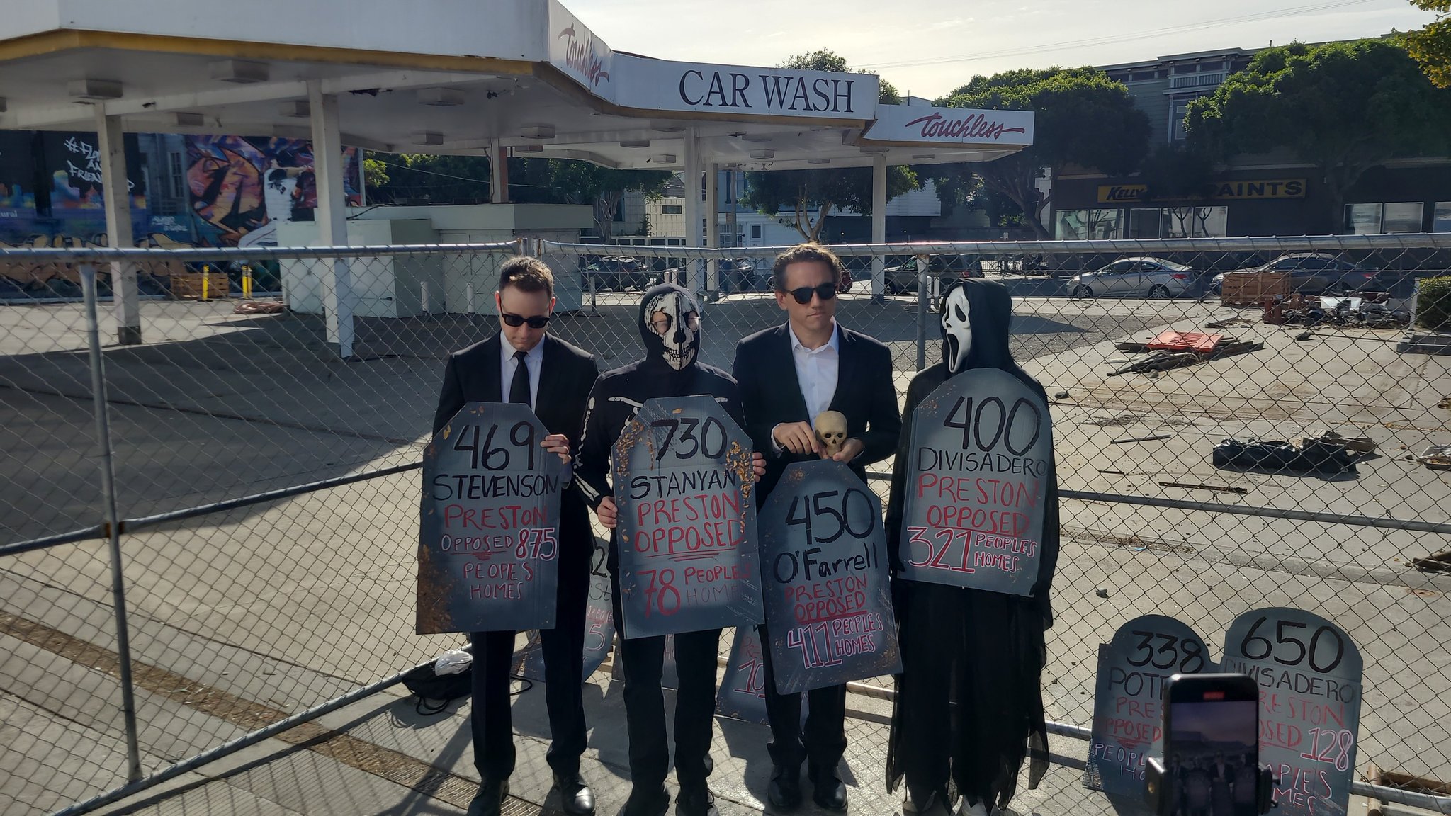 Four people in suits and masks stand holding signs resembling tombstones near a fenced construction site.