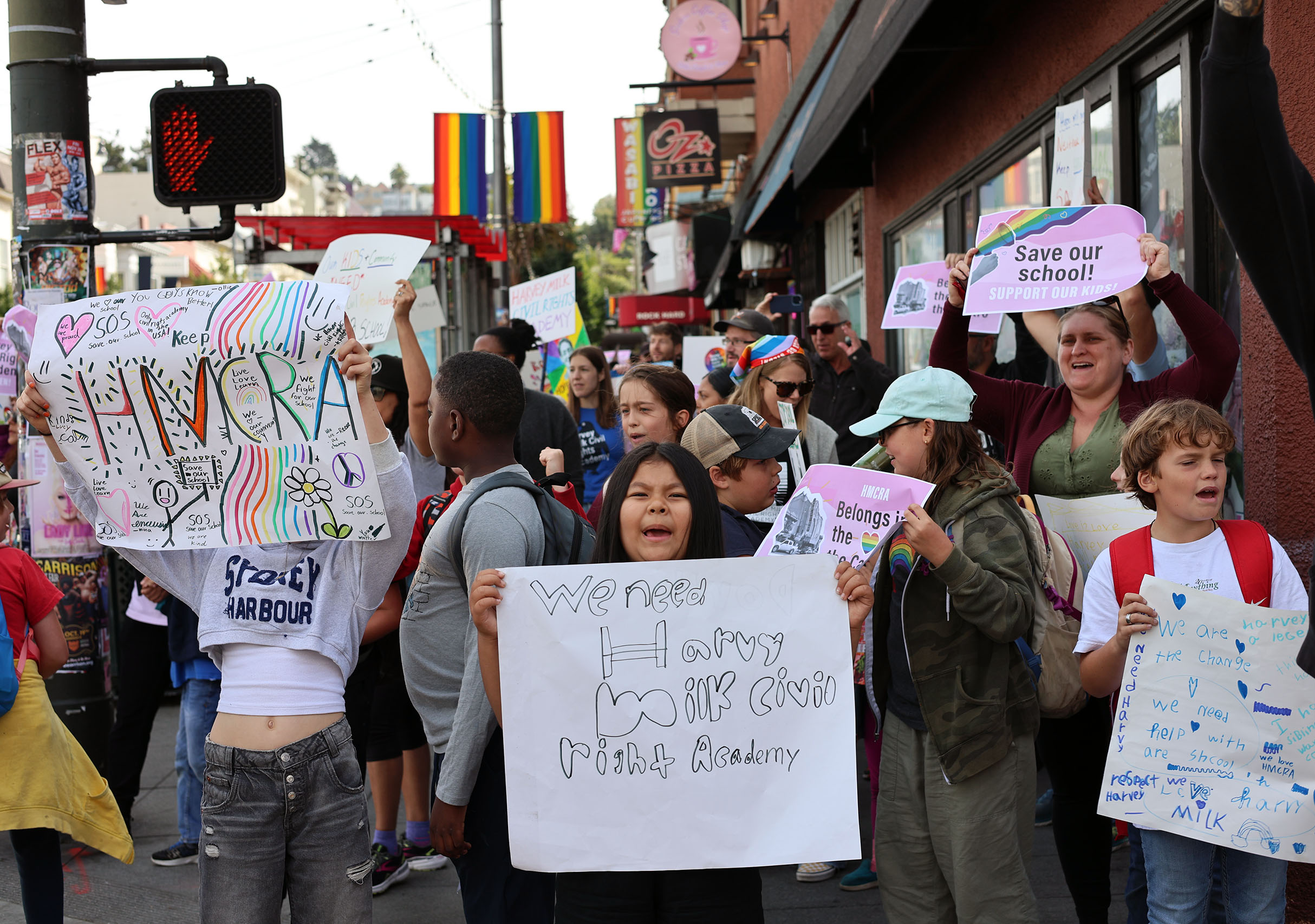 A group of people at a protest holding colorful signs with messages supporting LGBTQ rights, gathered on a street with a rainbow flag in the background.