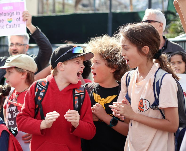 A group of children with backpacks are excitedly interacting outdoors, embodying the spirit of Harvey Milk. Some adults are visible in the background, one holding a sign.