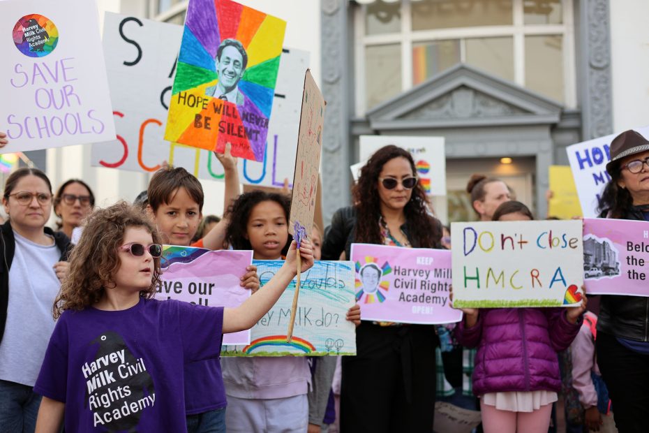 A group of people, including children, proudly hold signs advocating for the Harvey Milk Civil Rights Academy's mission.