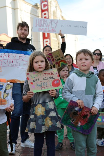 Children and adults hold protest signs advocating for their school, including a sign saying "Don't take our school away!" in an urban setting. The spirit of Harvey Milk echoes through the crowd, amplifying their resilience and determination to protect what they cherish.