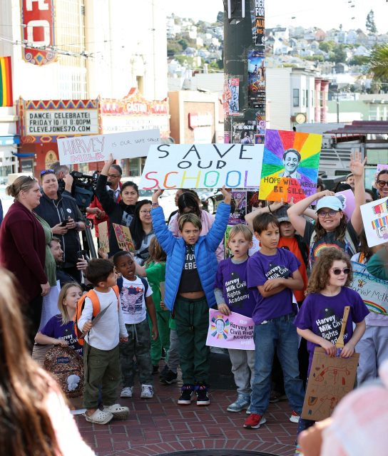 A diverse group of people, inspired by Harvey Milk's legacy, including children, hold colorful signs advocating for saving their school during a daytime rally in an urban setting.