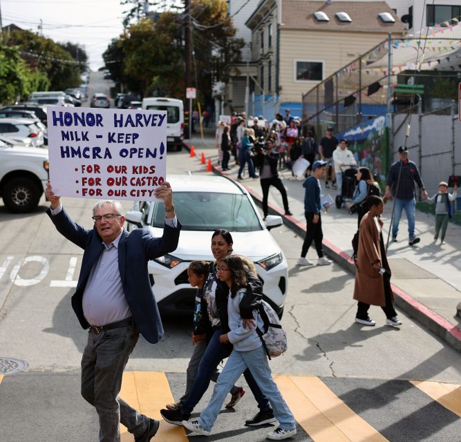Person holds a sign saying "Honor Harvey Milk - Keep HMCRA Open!" while walking across a street with others in an urban setting.