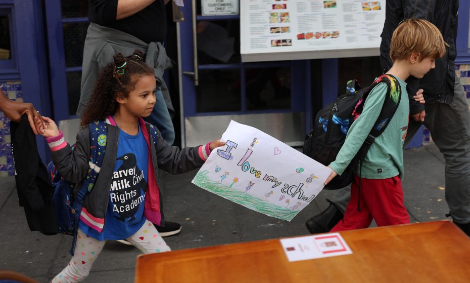 Two children holding hands, one proudly displaying a colorful drawing with "I love my school" written on it. They stroll joyfully near the blue building named after Harvey Milk, celebrating diversity and inclusion.