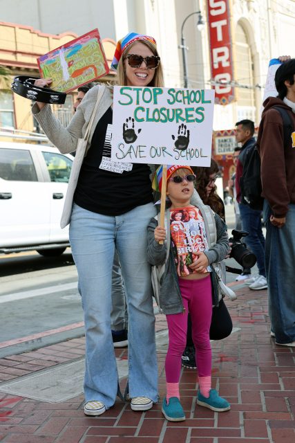An adult and a child hold signs protesting school closures on a bustling city street, channeling the spirit of Harvey Milk's activism.