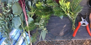 A variety of greenery and flowers lie on a wooden surface next to red-handled pruning shears, with a partially assembled wreath and a wire frame visible.