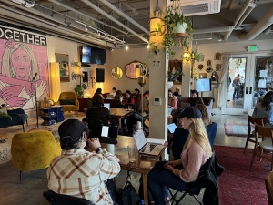 People are sitting and working on laptops inside a cozy cafe with plants and artwork on the walls.