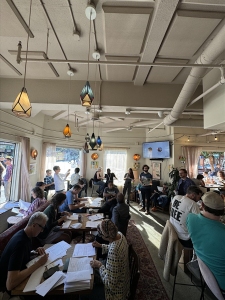 People are seated and standing in a bright café, engaged in discussion and paperwork. A TV screen is on the wall, and pendant lights hang from the ceiling.
