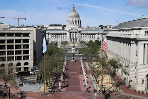 A cityscape view of a large domed building with an adjacent courtyard, lined with trees and flagpoles, under a clear blue sky.