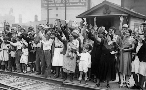 A crowd of people, including men, women, and children, stand by a railroad waving and cheering.