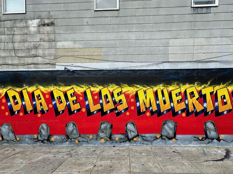 A vibrant mural features "Dia de los Muertos" in bold letters with gravestones and a stylized skeleton figure on a red and yellow background.
