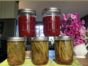 Five mason jars filled with preserved foods are displayed on a table. Three jars contain a red liquid, likely jam or preserves, while the other two are filled with green beans. A pink flower bouquet is in the background.
