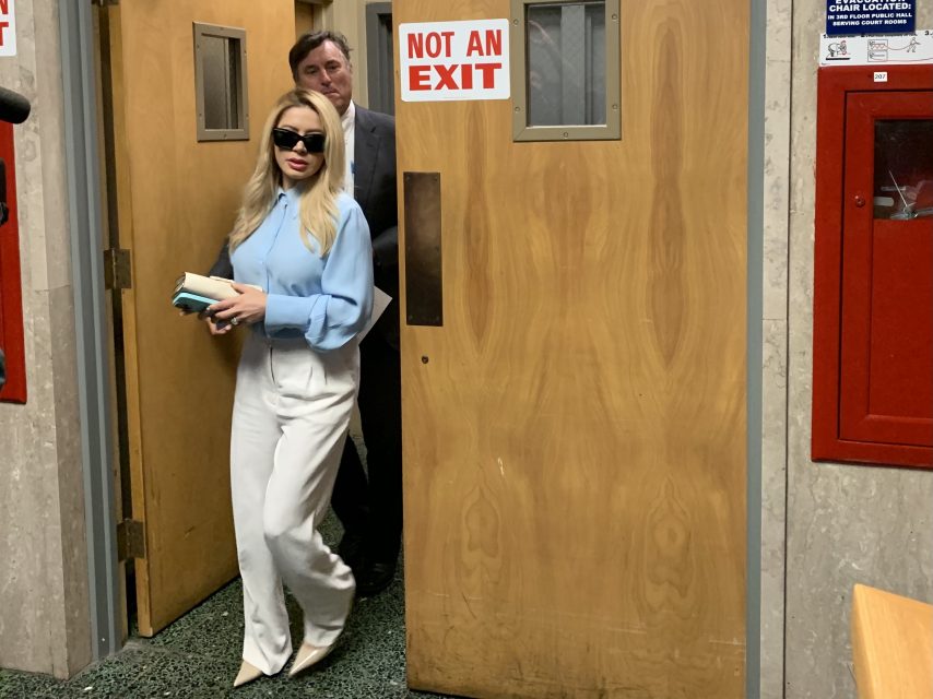 A woman in sunglasses and a blue blouse walks through a wooden door that says "Not an Exit," holding two books.