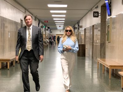 Two individuals walking down a corridor with benches and people in the background. The person on the left is in a suit, and the person on the right is wearing sunglasses and holding items.