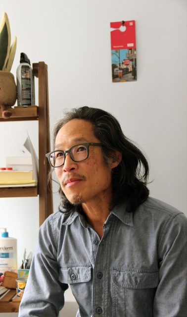 A person with glasses and long hair sits in a room with shelves, looking slightly upward.