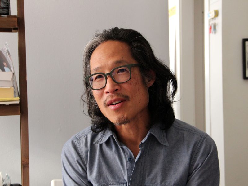 A person with long hair and glasses is sitting indoors, wearing a blue button-up shirt, with a neutral expression. Books are on a shelf beside them.