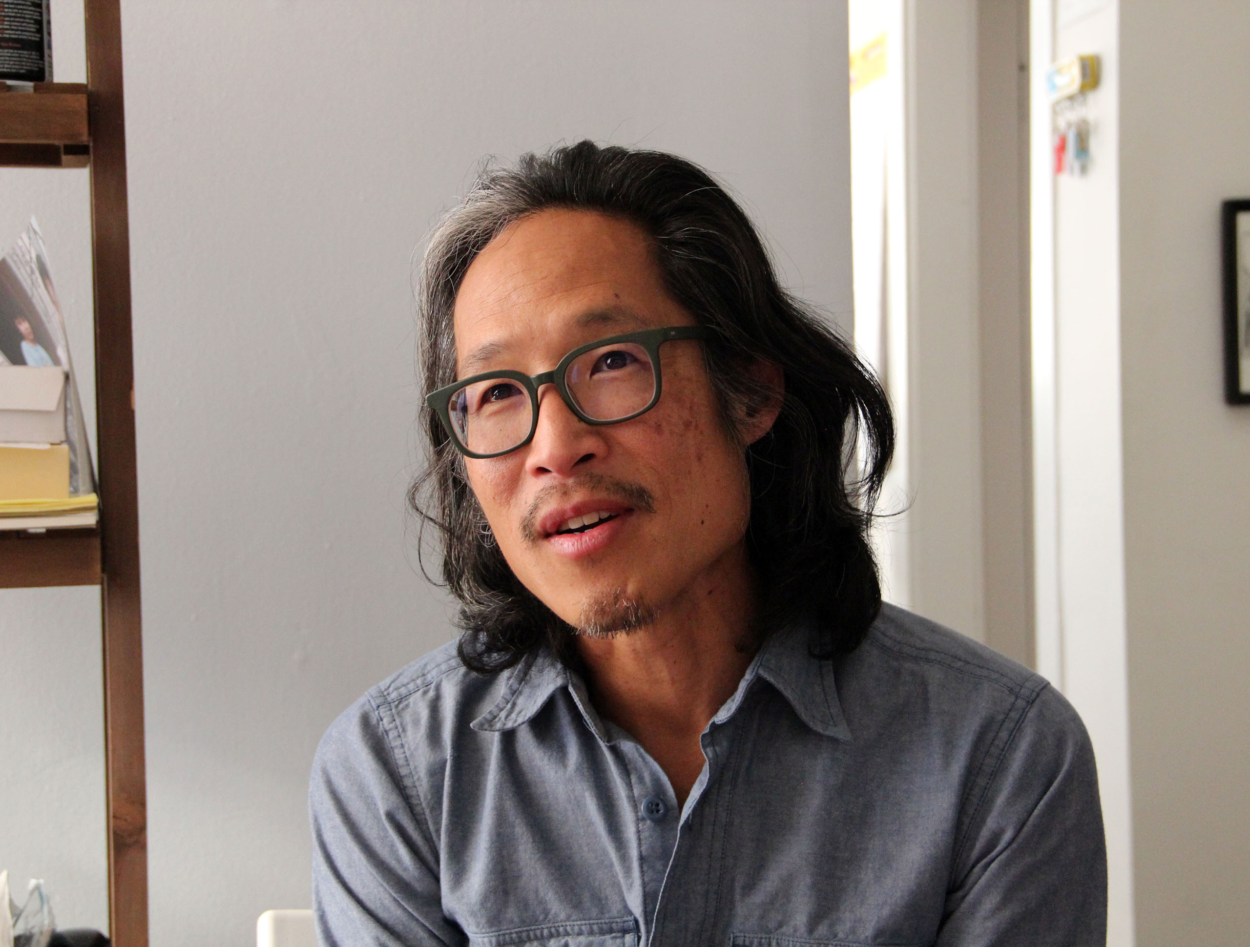 A person with long hair and glasses is sitting indoors, wearing a blue button-up shirt, with a neutral expression. Books are on a shelf beside them.