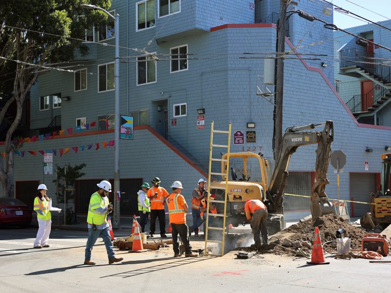 Construction workers wearing safety gear carefully operate machinery at a street excavation site, addressing a potential gas leak in front of a blue residential building.