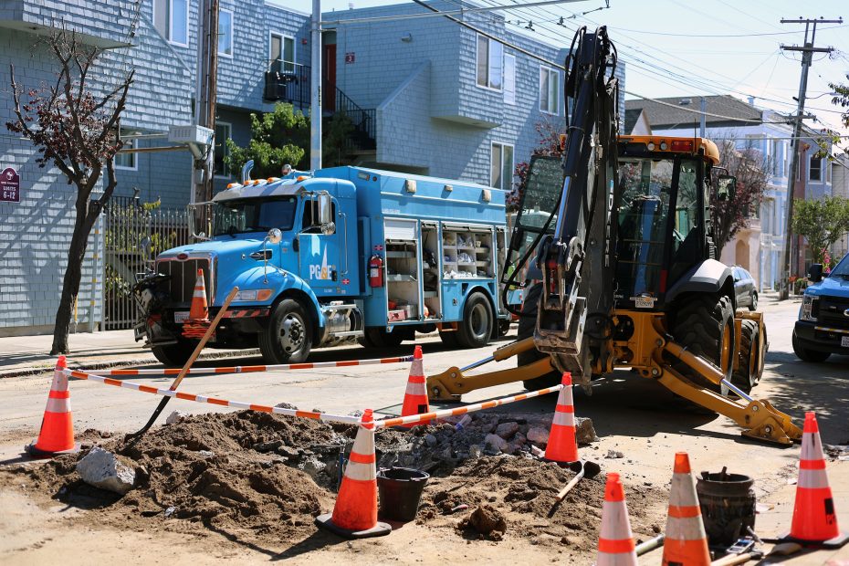 Construction site on a street with an excavator and utility truck working diligently to address a gas leak, surrounded by orange cones and piles of dirt.