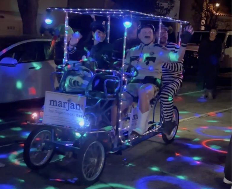 A group of people ride a pedal-powered vehicle adorned with vibrant lights in the night, showcasing a sign that reads "Marjan for Supervisor." Amidst the glow, the scene captures the spirit of an energetic SF election.
