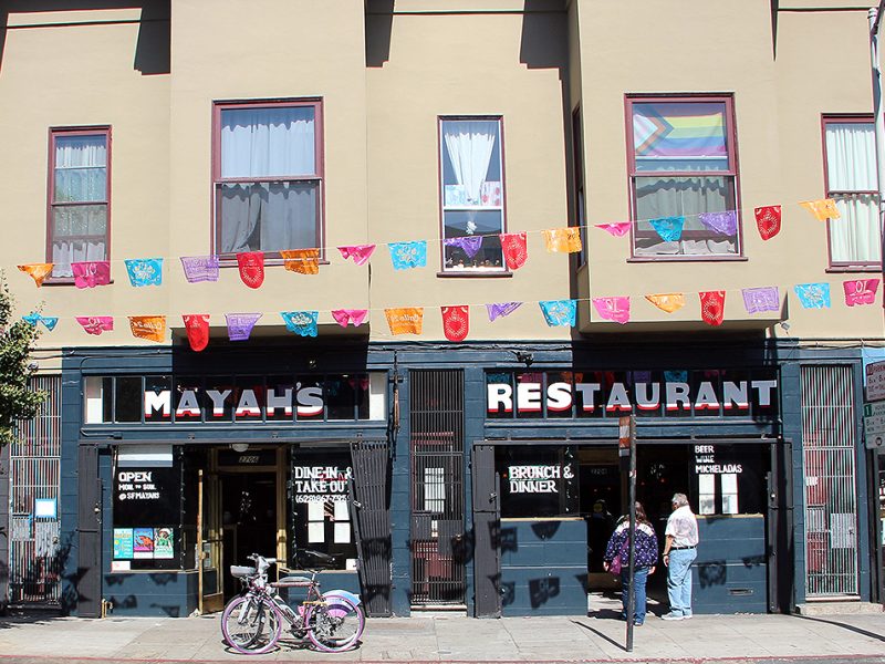 The street view captures Mayah's restaurant, adorned with a vibrant banner above. Bicycles are parked in front, and two people linger just outside the entrance, adding to the lively atmosphere.