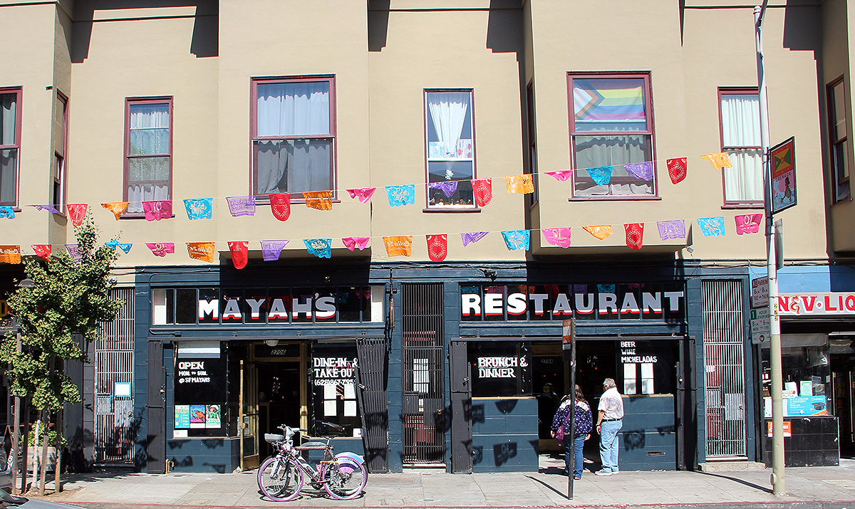 The street view captures Mayah's restaurant, adorned with a vibrant banner above. Bicycles are parked in front, and two people linger just outside the entrance, adding to the lively atmosphere.