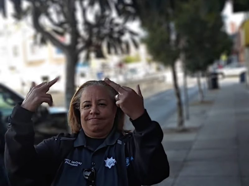A person in a police uniform is standing on a sidewalk, making a gesture with both hands. Trees and buildings are in the background.