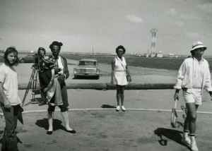 A black and white photo of four people on a road with a car in the background. One person carries a camera on a tripod. The setting appears to be open and rural.