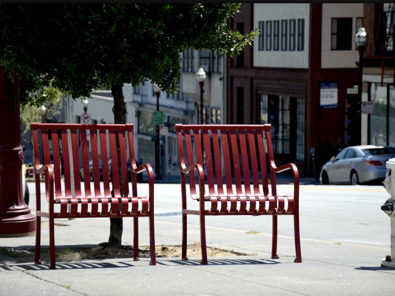 Two red metal benches face the street on a sidewalk next to a tree. Buildings and a parked car are in the background.